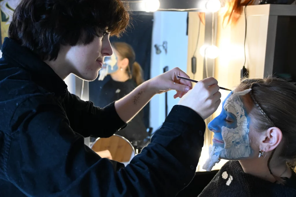 Math David Makeup applique du maquillage bleu et blanc sur le visage d'une jeune femme dans une loge.