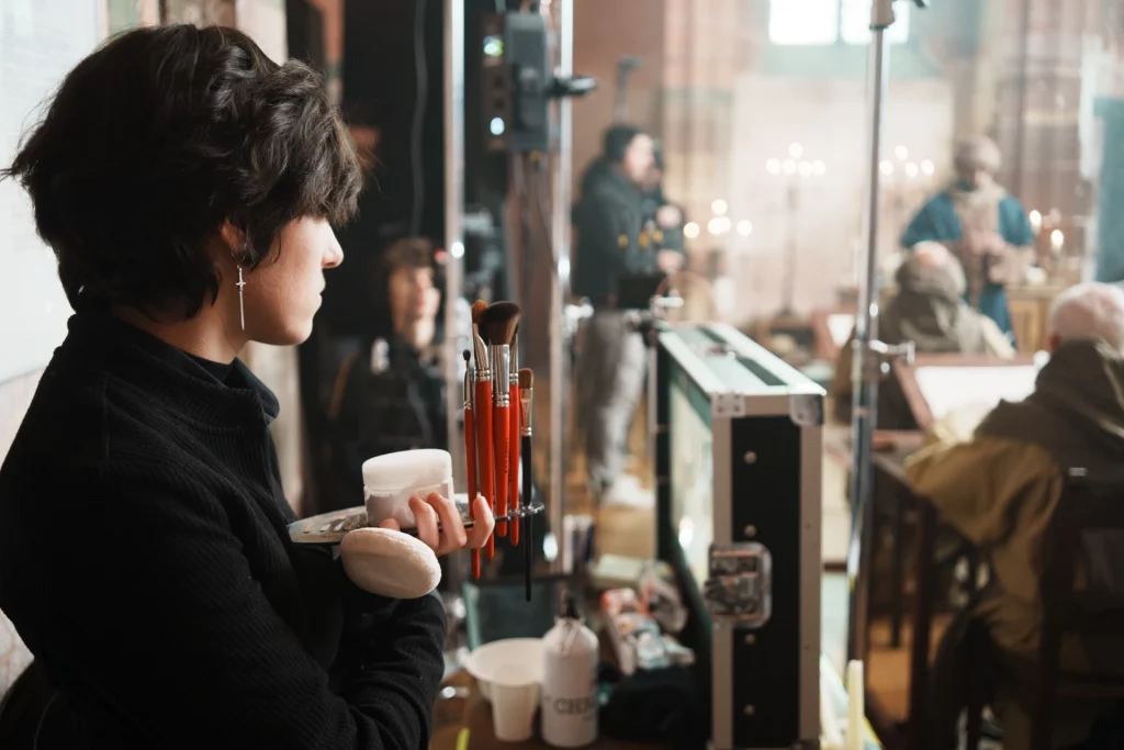 Math David Makeup sur un plateau de tournage, tenant des outils de maquillage, face aux acteurs en arrière-plan.