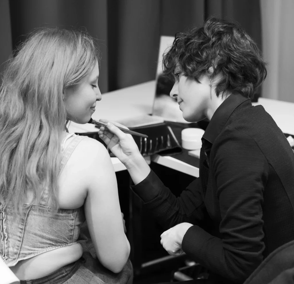 Math David Makeup qui maquille une jeune femme devant une table et un miroir.