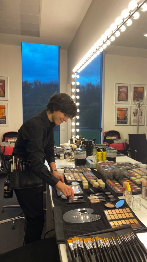 Math David Makeup dans les loges d'un tournage, devant son post de travail exposant son matériel, nettoie un pinceau.