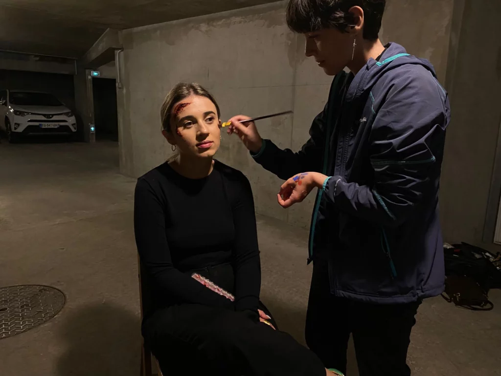Math David Makeup dans un parking pour un tournage, réalisant un maquillage SFX sur une jeune femme.