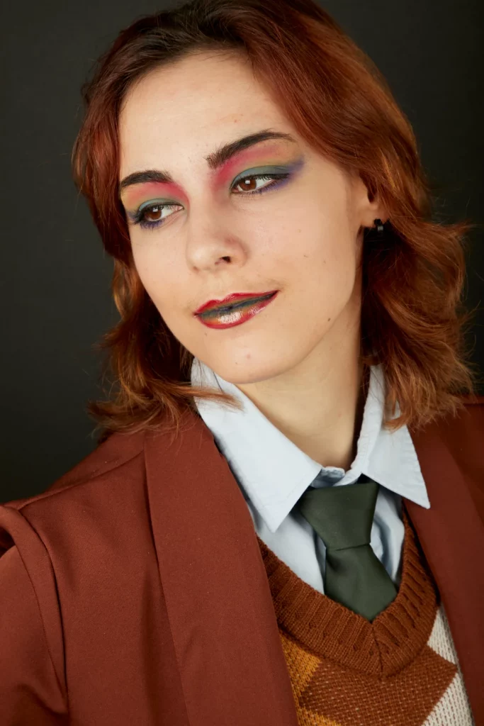 Portrait d'une femme au maquillage coloré, vêtue d'un blazer marron, d'un pull et d'une cravate.