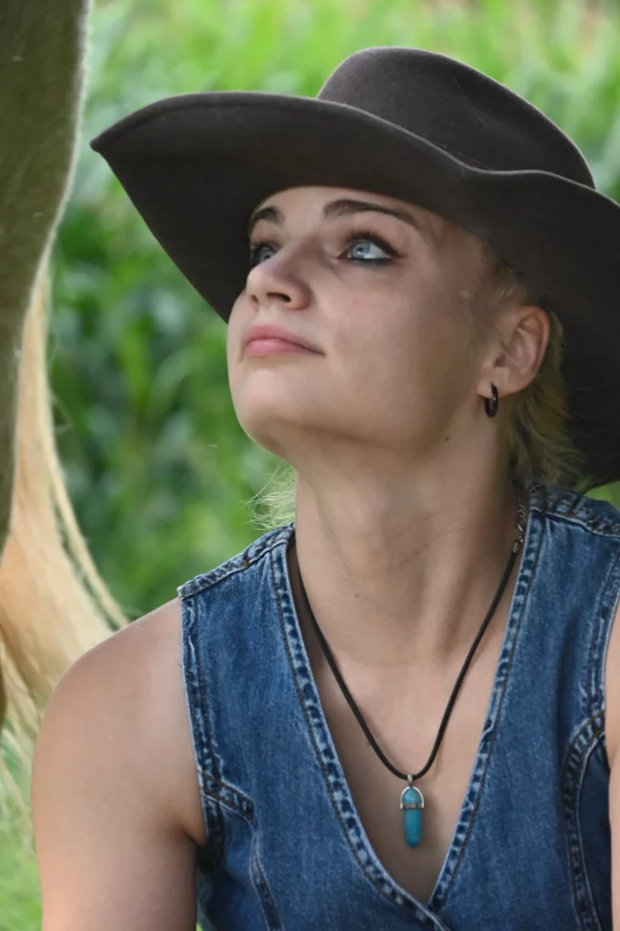 Une femme vêtue d'un gilet en jean et d'un chapeau à large bord lève les yeux, l'air pensif.