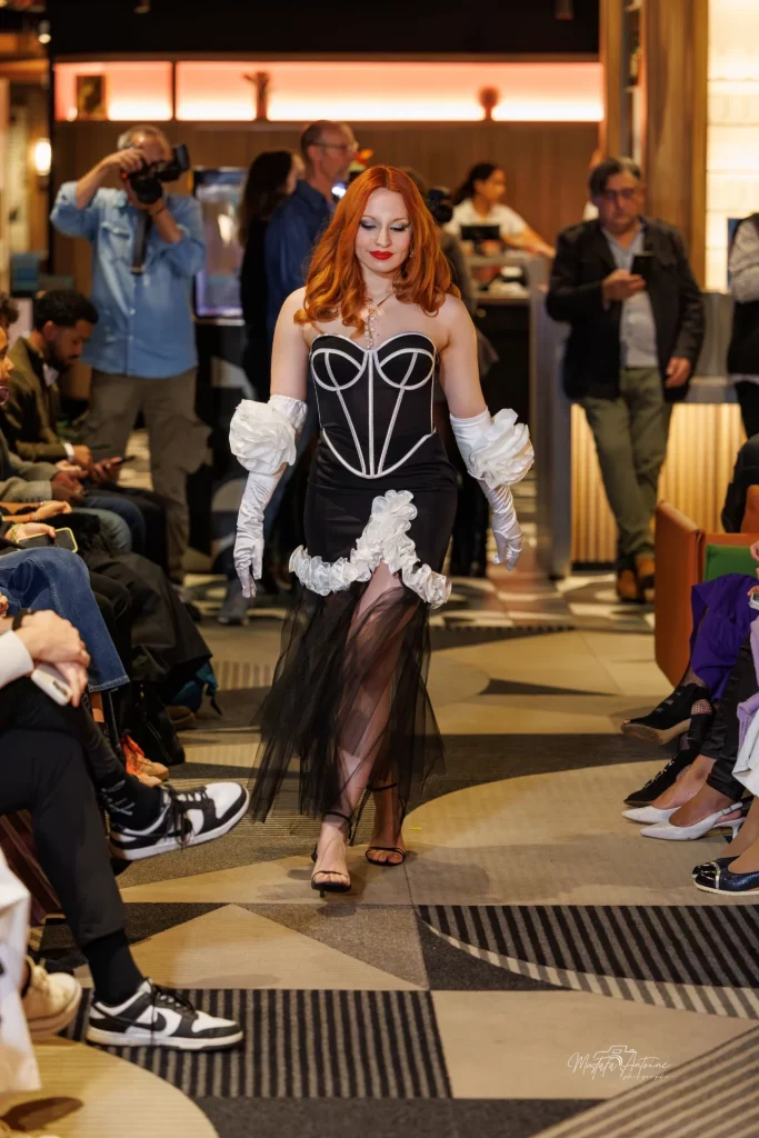 Une mannequin aux cheveux roux, vêtue d'une robe corset noire et de gants blancs, défile sur un podium.