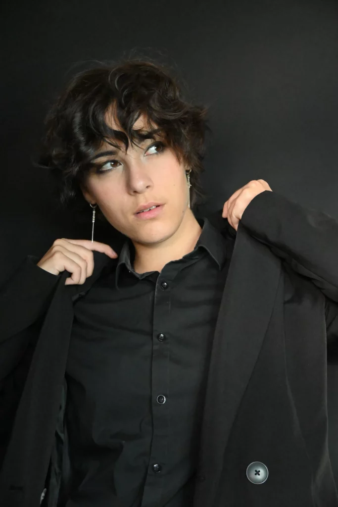 Femme aux cheveux bouclés, vêtue d'une chemise noire et d'une veste, avec un maquillage de jour sobre.