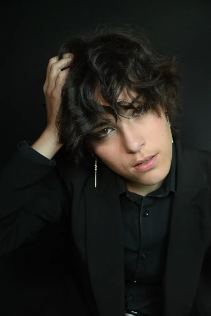 Une personne aux cheveux foncés et ondulés tient sa tête d'une main, vêtue d'un costume noir.