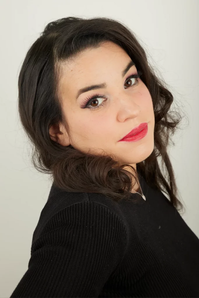 Une femme aux longs cheveux bruns et au rouge à lèvres rouge et d'un smoky eyes, vêtue d'un haut noir.