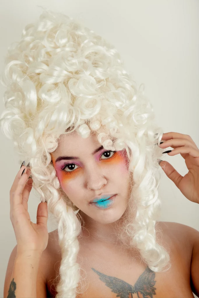 Une personne portant une grande perruque blanche bouclée, ombre à paupières rose et orange, lèvres bleues.