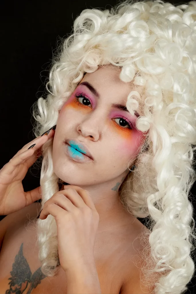 Femme portant une grande perruque bouclée, maquillée avec ombre à paupières rose et orange, lèvres bleues.