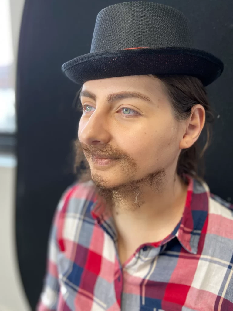 Modèle de profil avec une fausse barbe et un maquillage, portant un chapeau noir