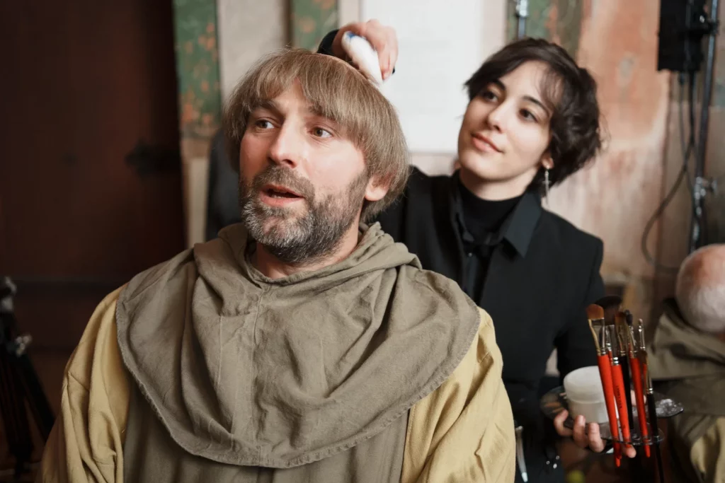 Homme déguisé en moine en premier plan avec derrière lui Math David Makeup qui fait des retouches maquillage.