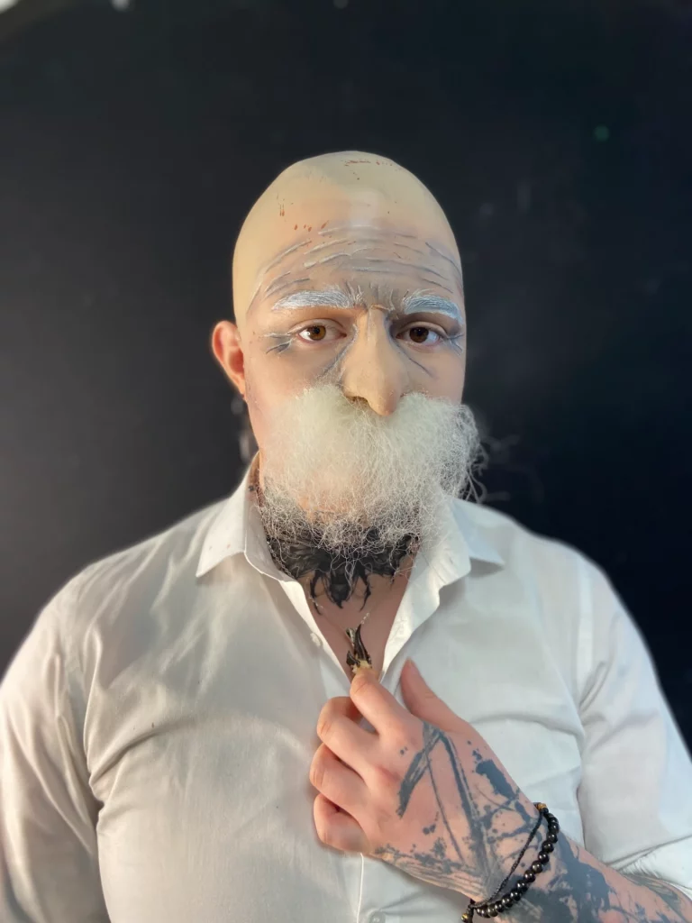 Une personne déguisée porte une calotte chauve et une barbe blanche touffue, vêtue d'une chemise blanche.