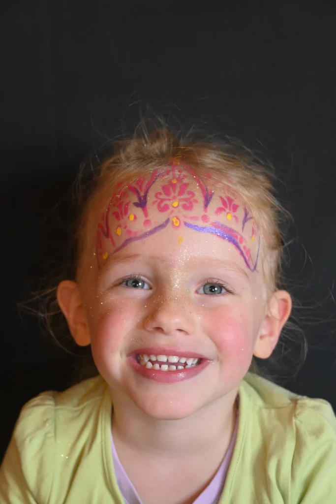 Jeune enfant souriante, le visage peint de couleurs vives et de motifs floraux ressemblant à une couronne.