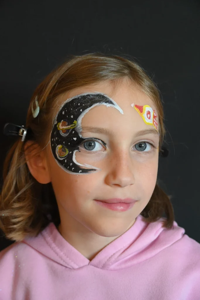 Jeune fille au visage peint de motifs célestes : une demi-lune et des planètes sur le côté droit, une fusée sur le côté gauche.