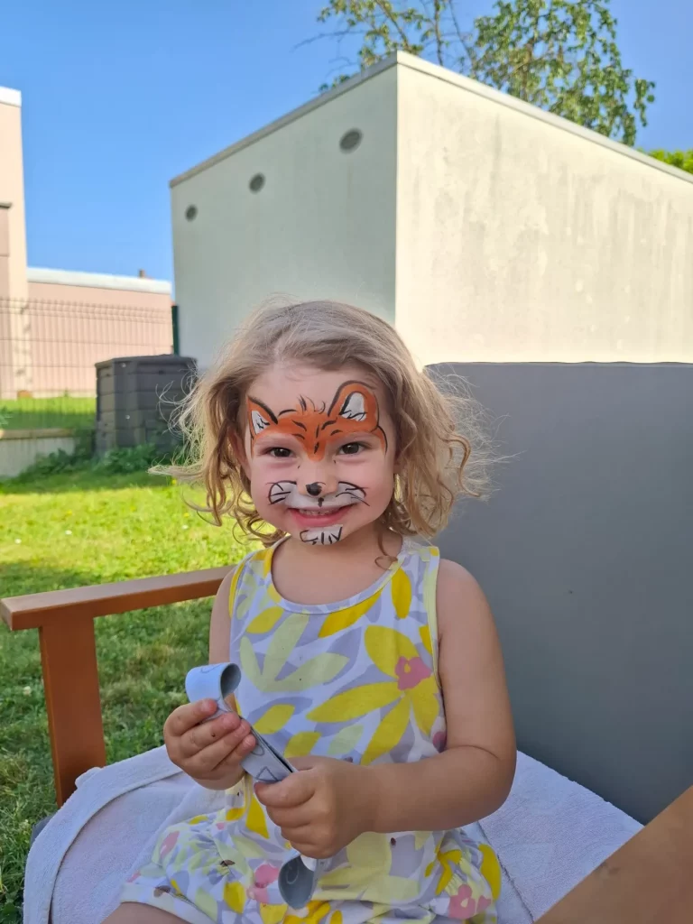 Une enfant assise et souriante aux cheveux bouclés et au visage peint en renard.