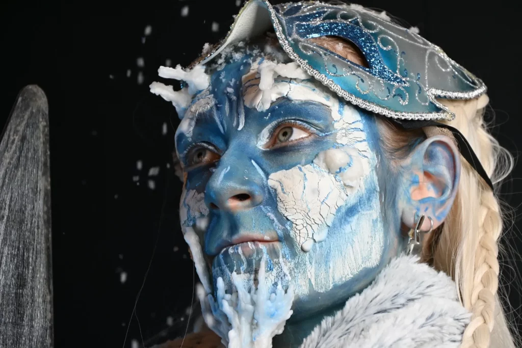 Une personne dont le visage est peint en bleu et blanc pour ressembler à de la glace, portant un masque argenté et orné.