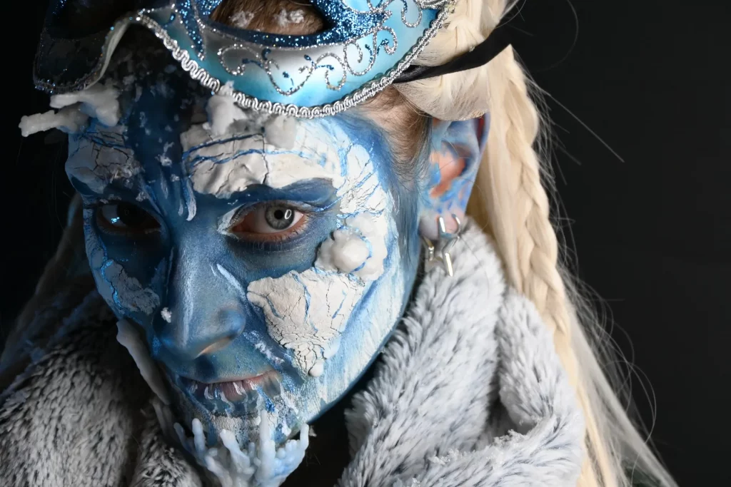 Femme au visage peint en bleu et effets de glace en relief, portant un masque de bal bleu un manteau de fourrure.