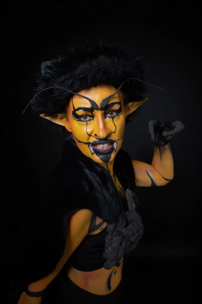 Femme avec un face et bodypainting illusion guêpe, aux oreilles pointues posant de manière théâtrale sur un fond sombre.