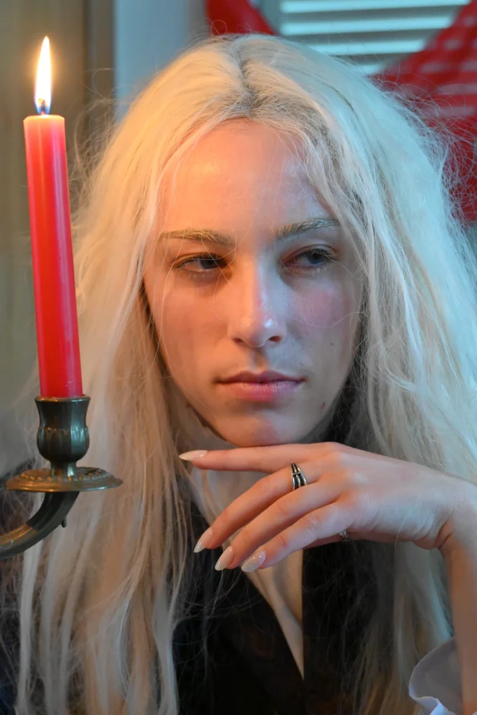 Une personne aux longs cheveux blonds pâles regarde pensivement au-delà d'une bougie rouge allumée.