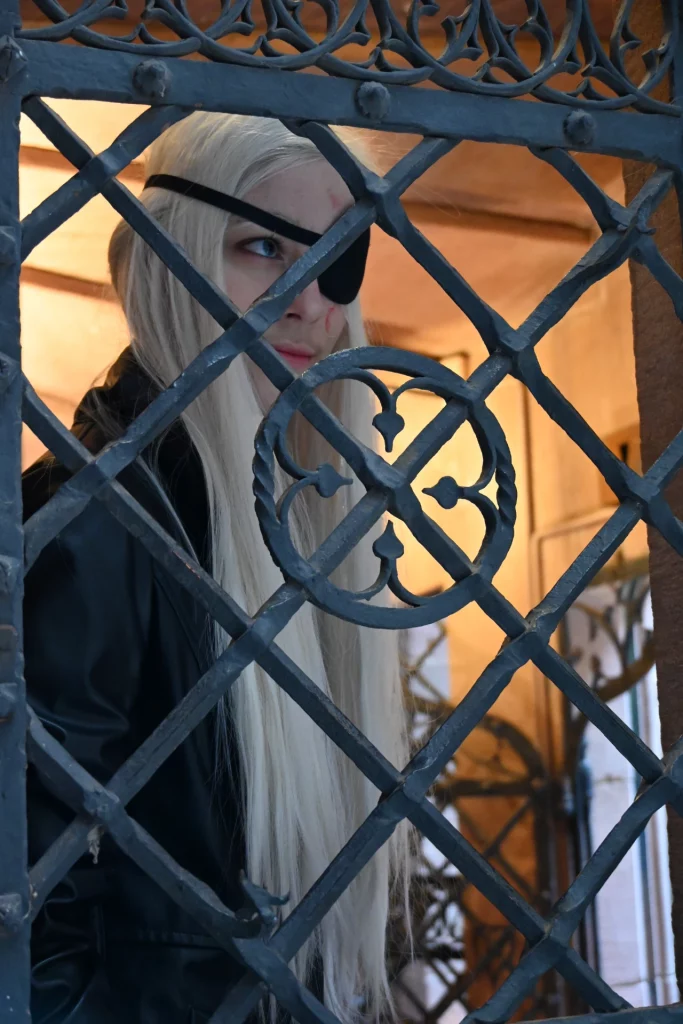 Une personne aux longs cheveux blancs et portant un cache-œil regarde à travers une grille en fer forgé.