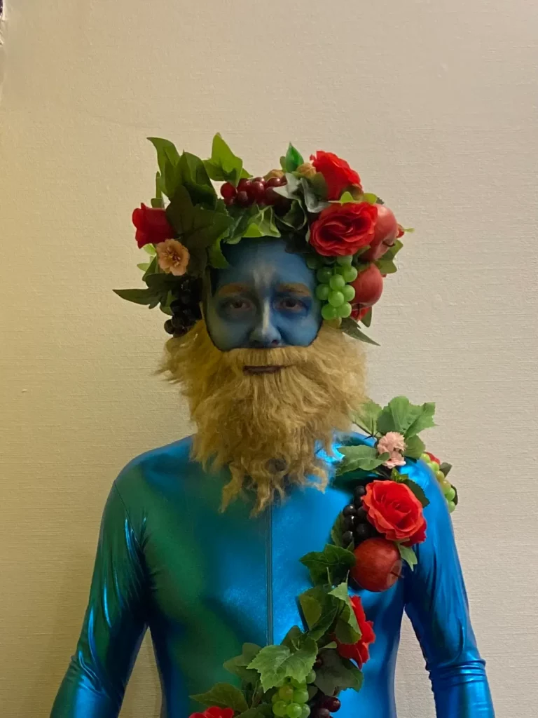 Une personne peinte en bleu pose dans un costume bleu brillant, orné d'une guirlande de roses rouges, de raisins et de verdure.