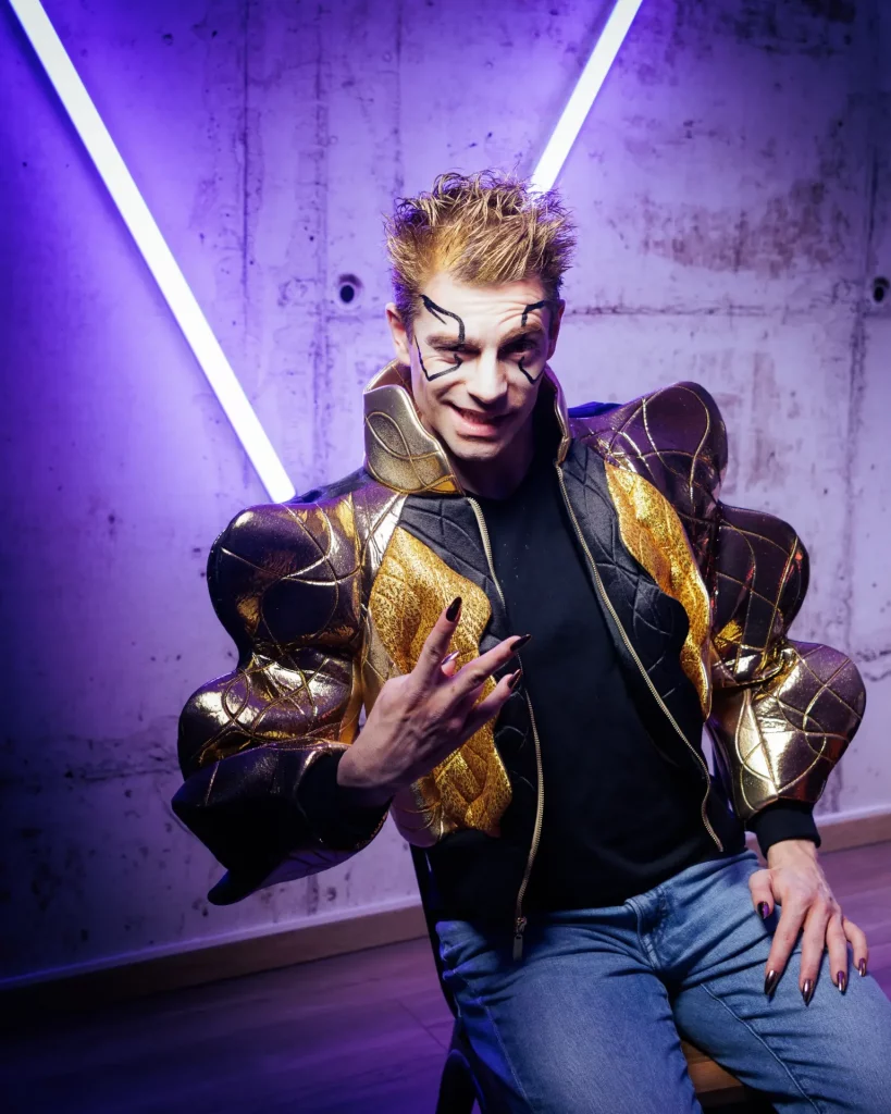 Une personne vêtue d'une tenue futuriste dorée et noire est assise contre un mur en béton éclairé par des néons violets.