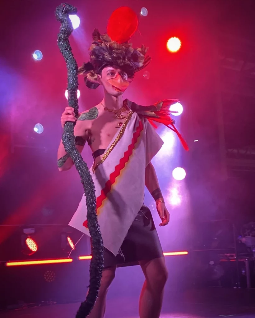 Une personne se tient sur scène dans un costume et maquillage de créature mythique type oiseau empereur.