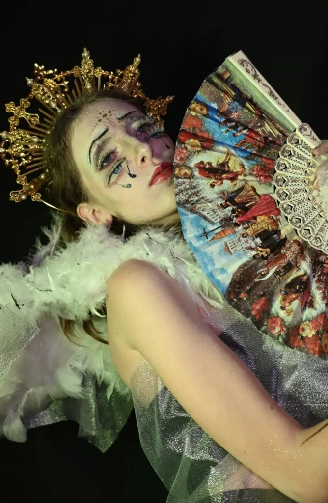 Une femme au maquillage théâtral et aux lèvres rouges, coiffée d'une couronne dorée tient un éventail coloré et orné.